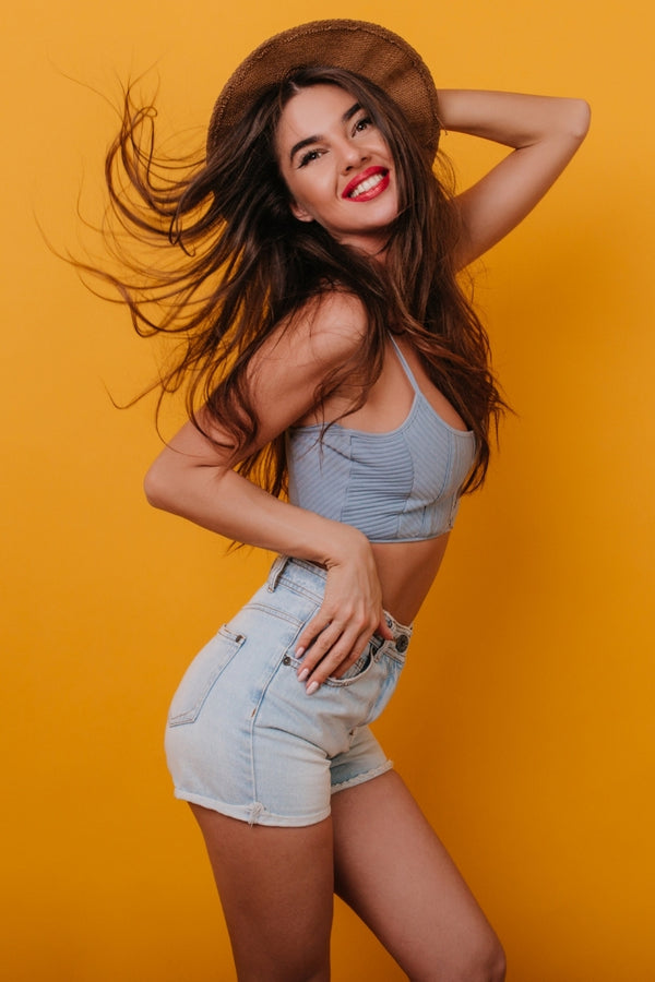 Glowmedik Model in a light blue crop top and denim shorts with a wide-brimmed hat against an orange background