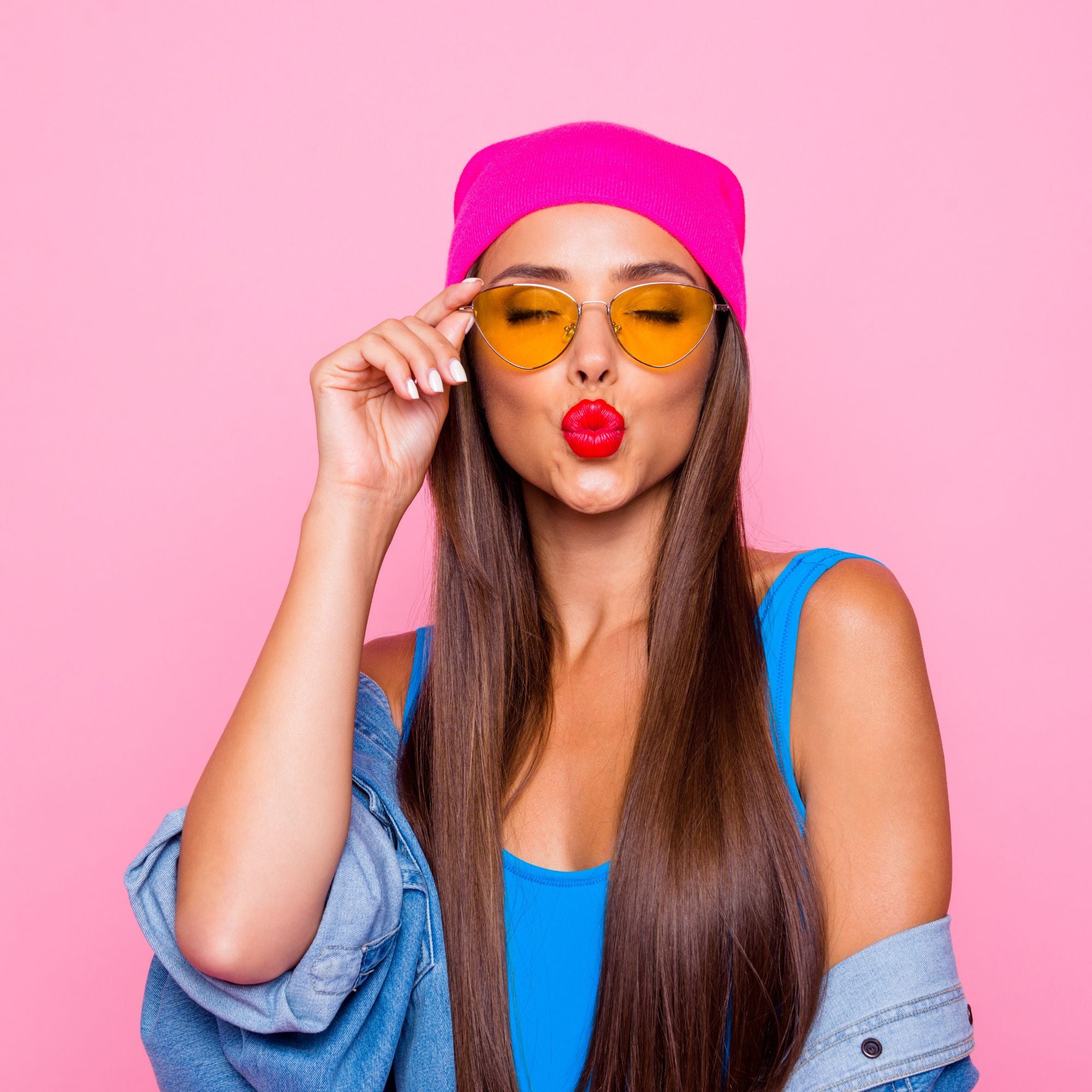 Woman wearing a pink cap, blue top, and yellow sunglasses on a pink background